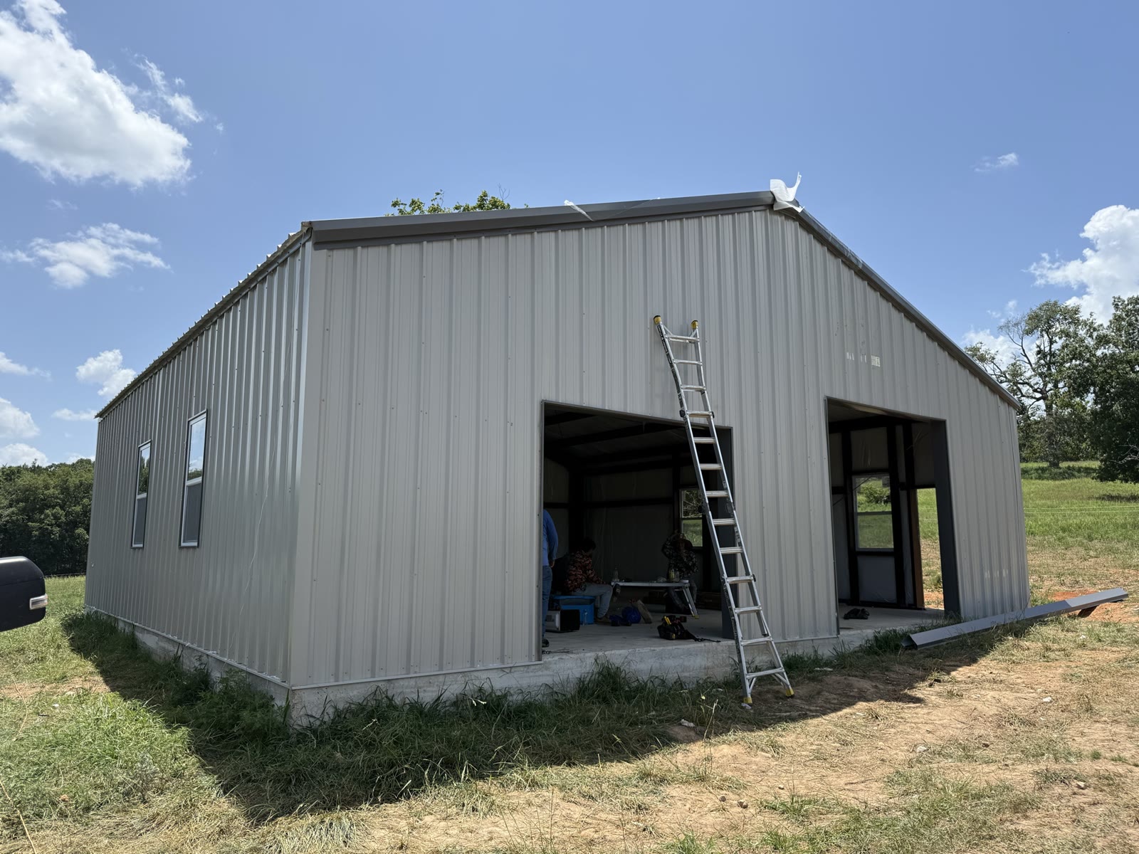 Steel workshop with light gray wall panels and roof installed, two large door openings still open, an extension ladder leaning against the wall and crew working inside on the concrete slab.