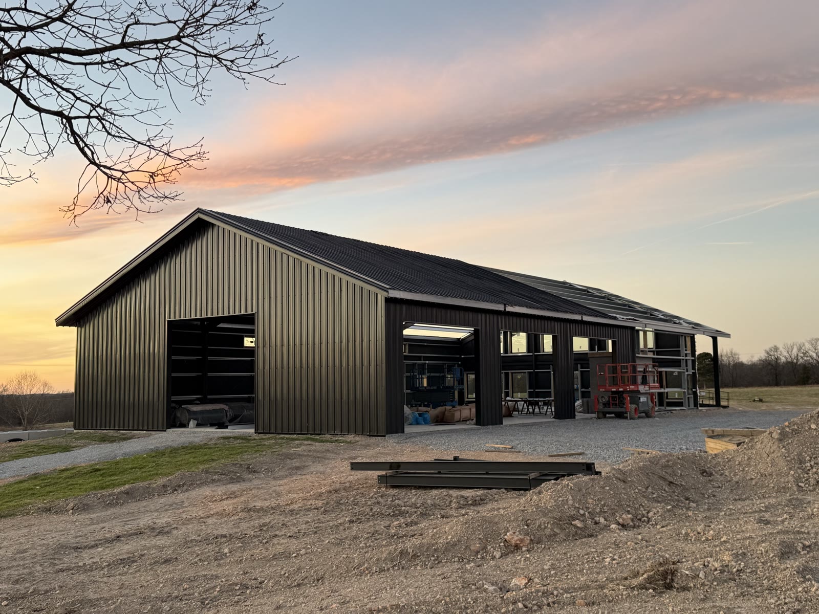 Dark metal-clad steel barndominium at sunset with the roof panels and wall skin installed, window openings and a large door opening still to be filled, gravel drive in the foreground.