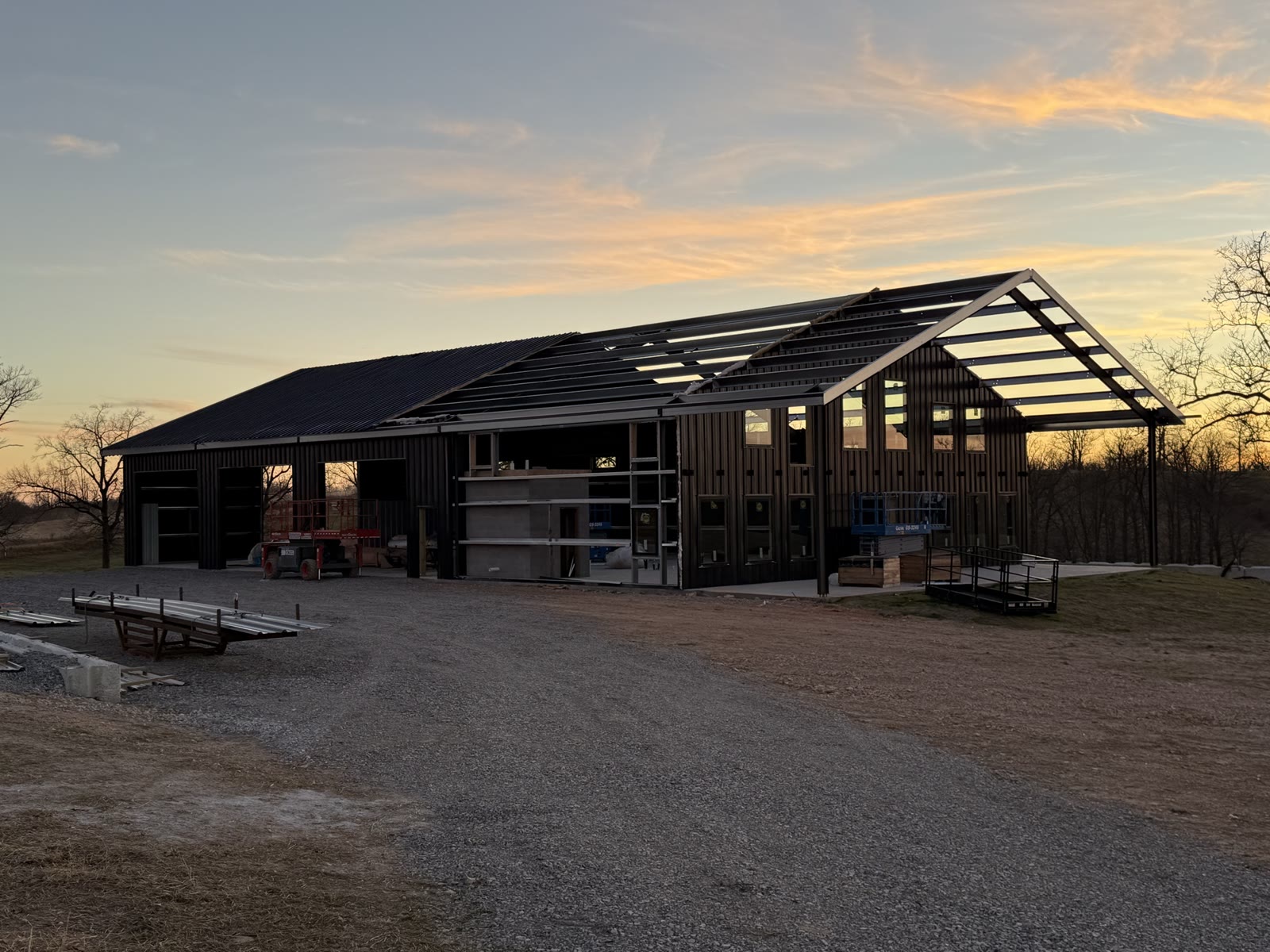 Long steel-frame building at dusk with roof panels and most wall panels installed, an open porch bay at the gable end still bare frame, gravel drive and loose steel stacked in front.