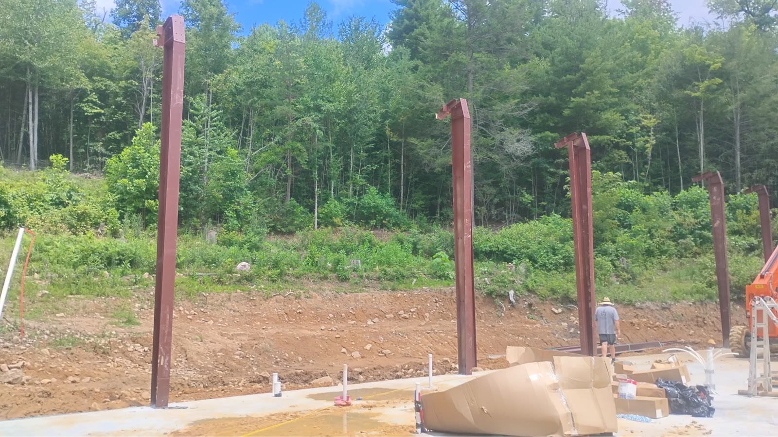 Red-iron columns standing anchored to a fresh concrete slab on a Tennessee hillside site, with a crew member and a lift working at the far end and bare cut earth behind the pad.
