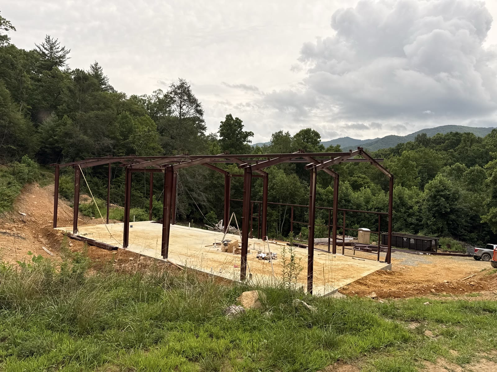 Red-iron steel frame erected on a poured slab cut into a wooded Tennessee hillside, with rafters and columns set, temporary cable bracing in place, and forested mountain ridges in the background.
