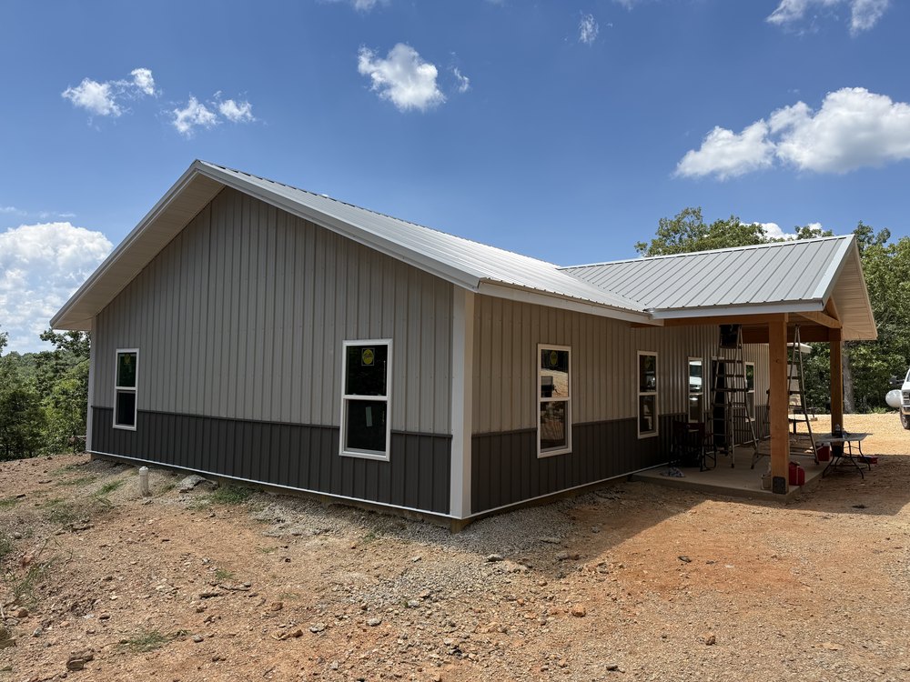 Completed Greers Ferry barndominium, front elevation with cedar timber-column gable porch, metal roof, and two-tone ash gray and charcoal wainscot panels.
